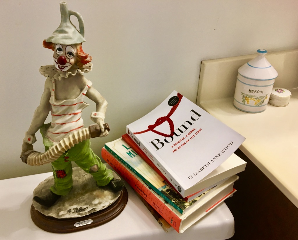 a stack of books on the tank of a toilet. The top book is Bound: A Daughter, a Domme, and an End-of-Life story. Beside the books is a ceramic clown.